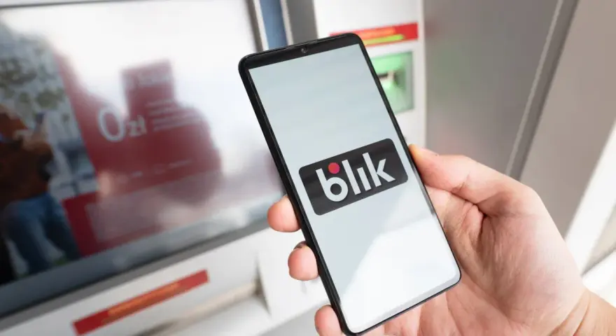 Dłoń trzyma smartfon z logo BLIK przed bankomatem.
