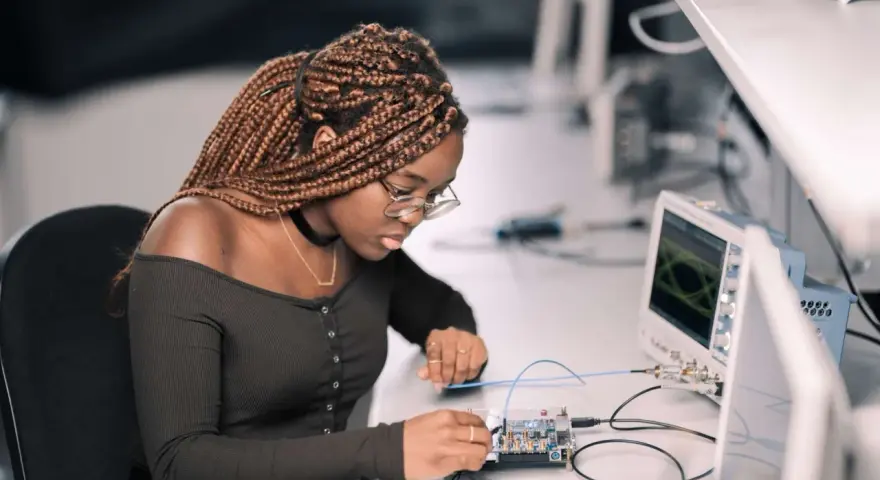 Junge Frau mit geflochtenen Haaren arbeitet konzentriert an einer Platine, während ein Oszilloskop im Hintergrund läuft. Ein spannendes elektrotechnik studium.