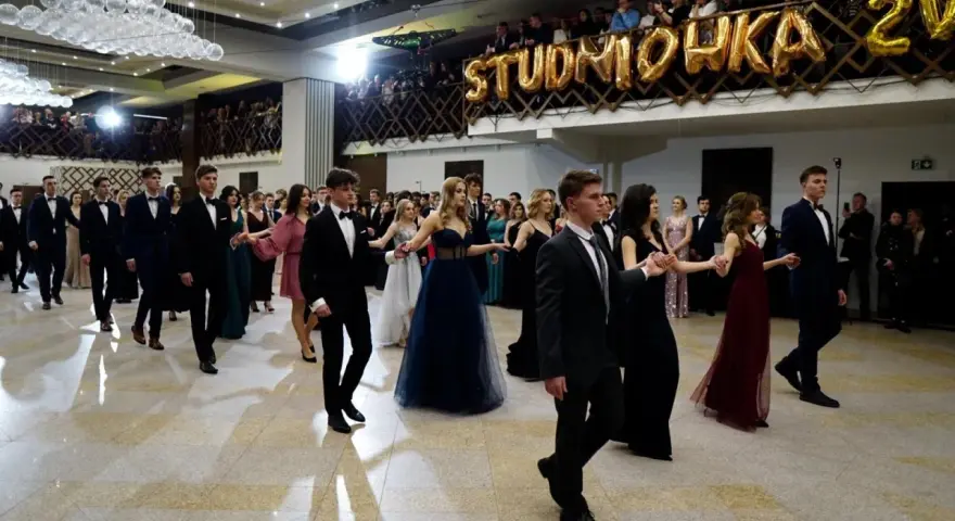 Młodzi ludzie w eleganckich strojach, tańczący poloneza na balu studniówkowym.