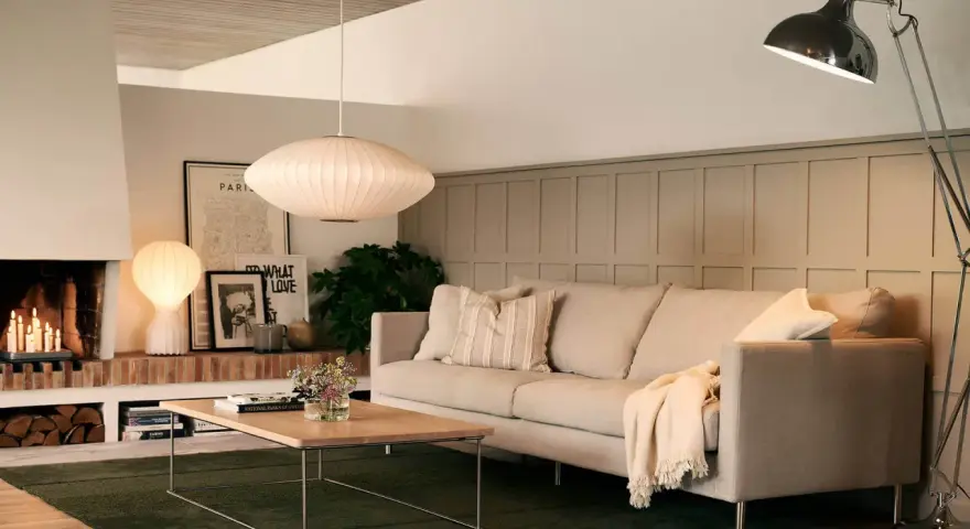 En ljus soffa med kuddar och en filt, framför ett soffbord i trä. En golvlampa och en taklampa lyser upp rummet. Perfekt för att mäta soffa mått.