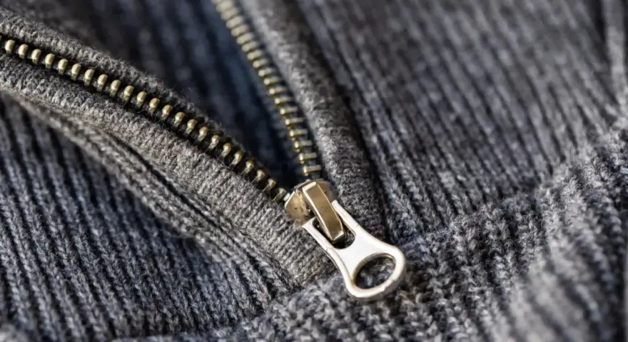 Metalowy suwak na szarym, dzianinowym materiale. Zamek jest częściowo rozpięty, co pokazuje, w którą stronę otwiera się zamek.