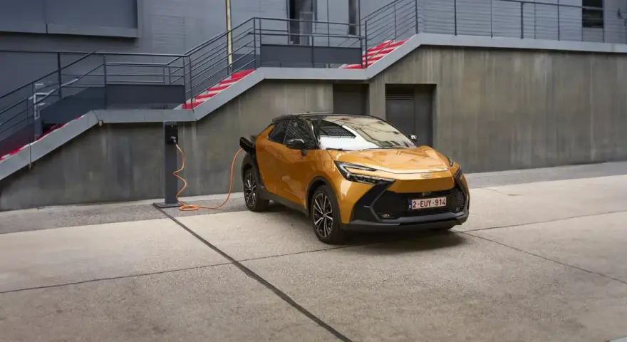 Złoty Toyota plug-in z pomarańczowym kablem ładowania podłączonym do stacji. Nowoczesna architektura w tle.