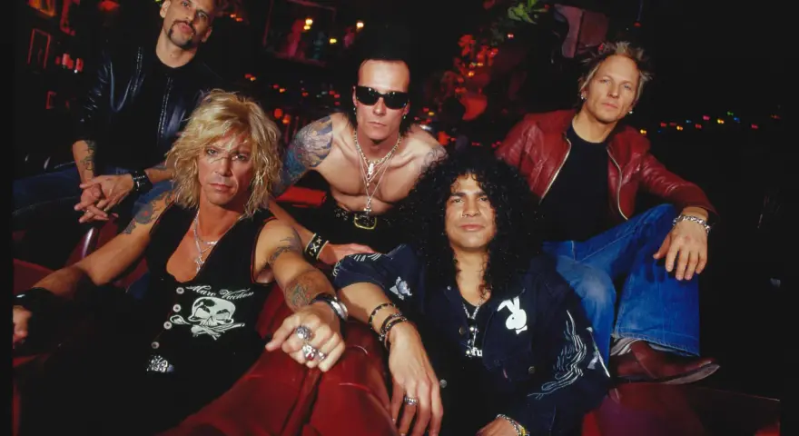 Cinq musiciens, un groupe de slash, posent sur un canapé rouge. Ils portent des vestes en cuir et des bijoux.