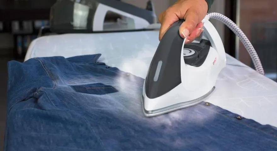 Hand using a steam iron on a denim shirt on an ironing board.