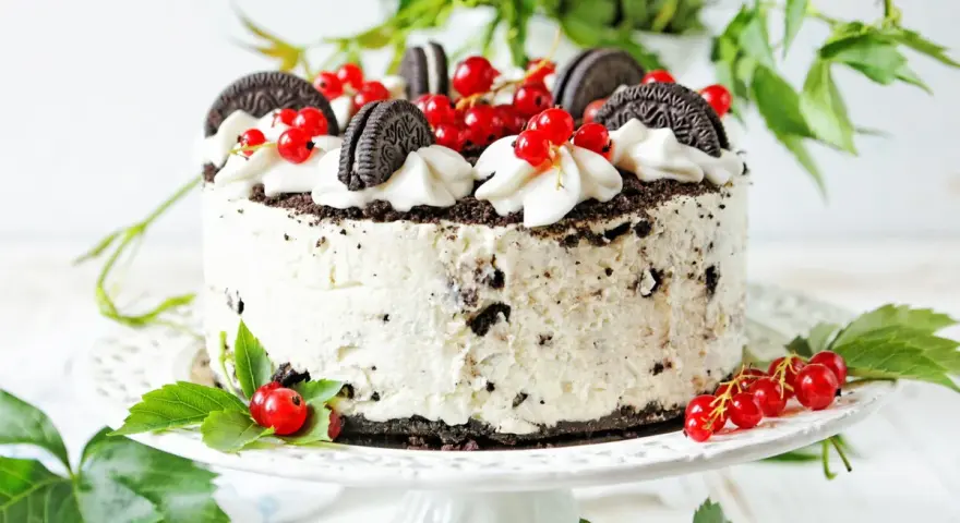 Pyszny tort z kawałkami oreo ciastka, udekorowany bitą śmietaną i czerwonymi porzeczkami.