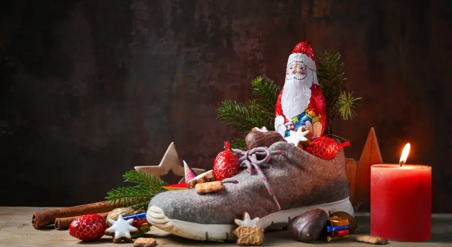 Ein Schuh gefüllt mit Leckereien, daneben ein Schokoladen-Nikolaus, Tannenzweige und eine brennende Kerze.