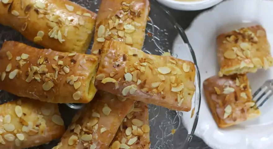 Omas Apfeltaschen Rezept: goldbraune, glasierte Gebäckstücke mit Mandelblättchen, serviert auf einem Glasständer und einem Teller, daneben eine Tasse Kaffee.