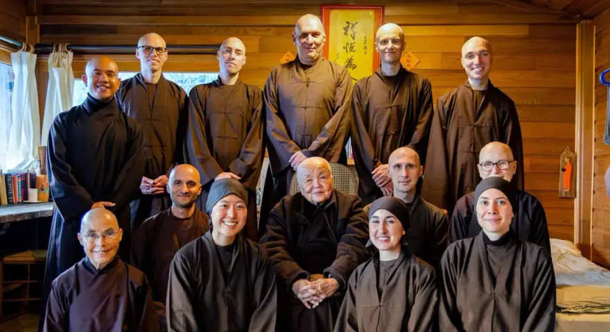 A group of monks and nuns, including an elder monk person in the center, pose for a photo in a room with wooden walls.