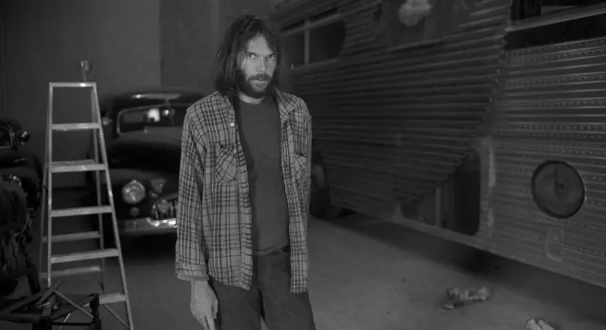 Neil Young, jung und nachdenklich, steht vor einem umgebauten Bus. Eine Leiter und ein Oldtimer im Hintergrund.