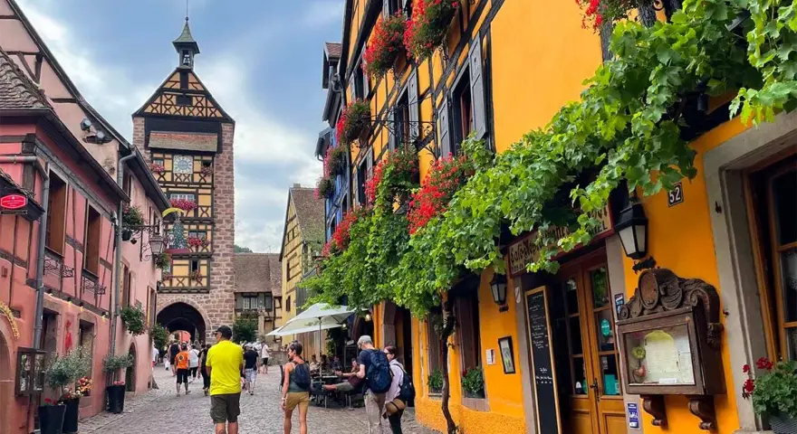 Cobblestone street in a picturesque Alsace town, with colorful half-timbered buildings adorned with flowers. A clock tower stands tall in the background.