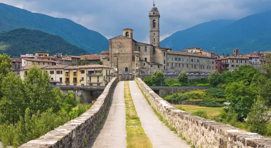 Ponte medievale in pietra che conduce a un borgo con chiesa e campanile, incastonato tra le verdi montagne degli Appennini Emilia Romagna.