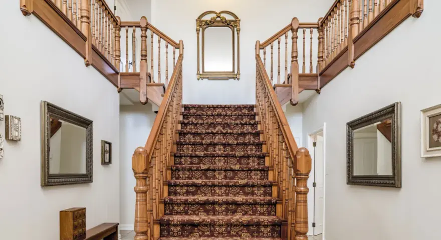 Elegante escalera antigua con alfombra de estilo persa y barandillas de madera. Un espejo dorado decora la pared central, complementando el revestir escalera antigua.