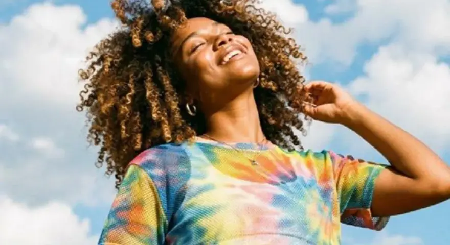 Une femme souriante aux cheveux bouclés, sous un ciel bleu nuageux. Elle porte un t-shirt tie-dye coloré. Une belle image pour apprendre comment rendre une photo flou.