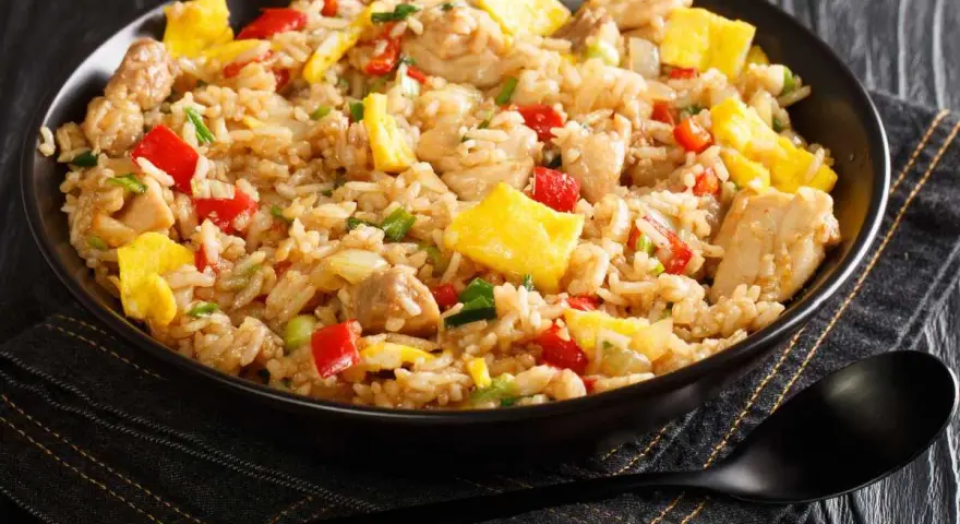 Un plato humeante de arroz chaufa con trozos de pollo, huevo revuelto, pimientos rojos y cebolla verde.