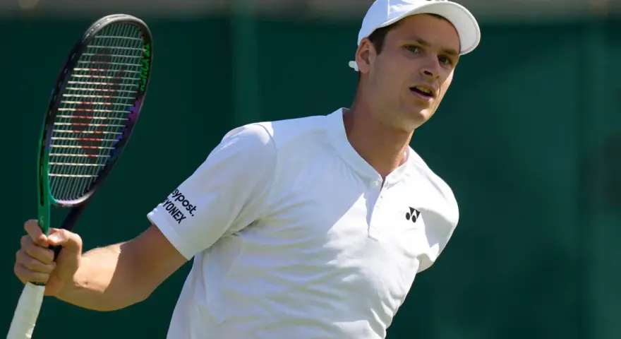 Hubert Hurkacz w białej koszulce i czapce, gotowy do uderzenia rakietą na Wimbledonie.