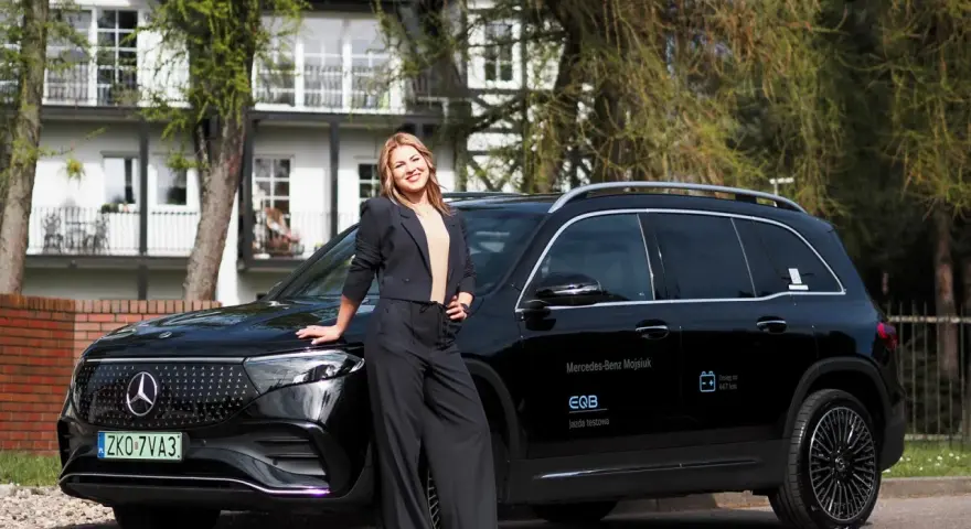 Kobieta w czarnym garniturze opiera się o czarnego Mercedesa EQB na tle białego domu.