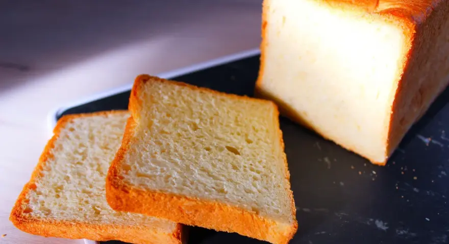 Pain de mie moelleux, parfait pour une recette pain de mie boulangerie maison. Deux tranches dorées et un pain entier sur un plateau sombre.