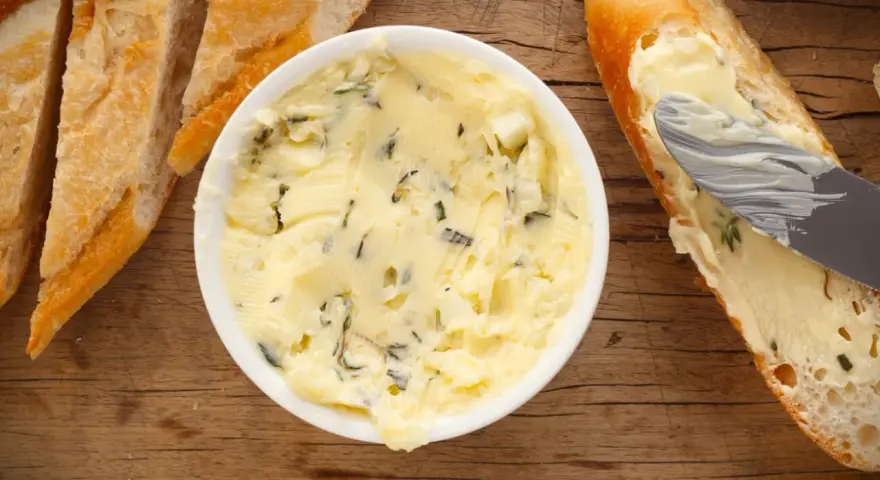 Knoblauchbutter selber machen ist einfach! Hier ist eine Schüssel mit selbstgemachter Knoblauchbutter, daneben Baguettescheiben und ein Messer, das Butter auf ein Brot streicht.