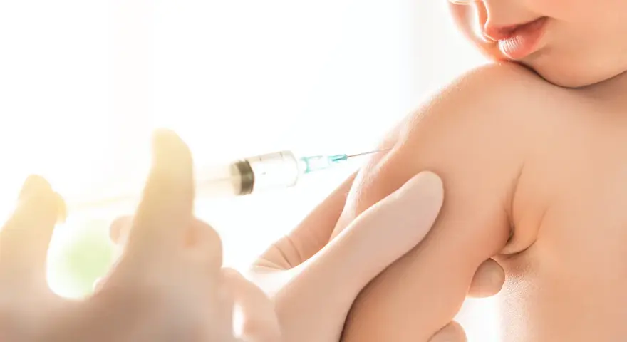 Dziecko otrzymuje szczepionkę 6w1. Ręka w rękawiczce trzyma strzykawkę z igłą, podając zastrzyk w ramię malucha.
