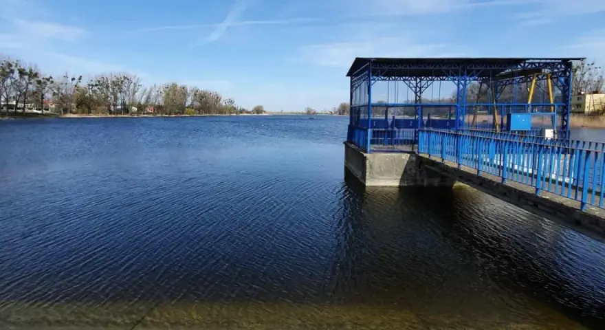 Spokojne jezioro średzkie z błękitną budowlą i pomostem. W tle drzewa i zabudowania.