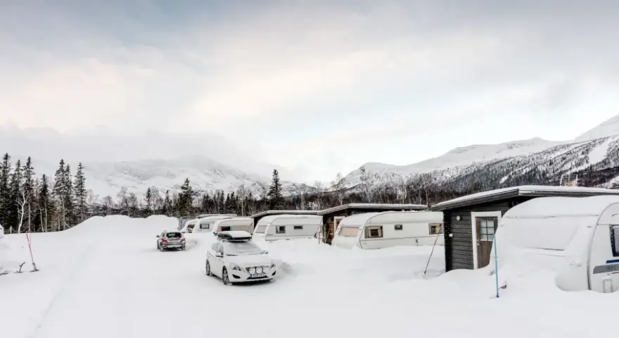 Vinterlandskap vid moskosel camping med snötäckta husvagnar och bilar, berg i bakgrunden.