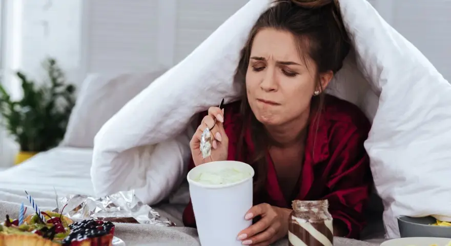Ragazza sotto le coperte mangia gelato, dolci e pasta. La sua espressione suggerisce una forte fame pre ciclo.