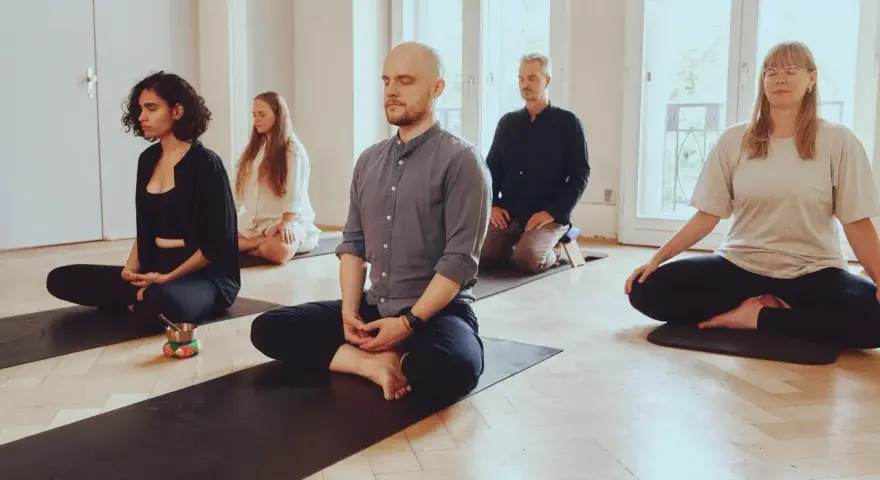 Menschen in einem hellen Raum meditieren auf Matten. Die Meditation wirkt beruhigend und fördert die Konzentration.