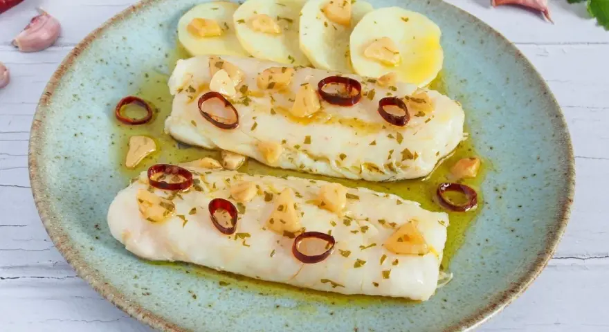 Dos filetes de merluza a la bilbaina, con patatas, ajo laminado y guindilla, en un plato azul.