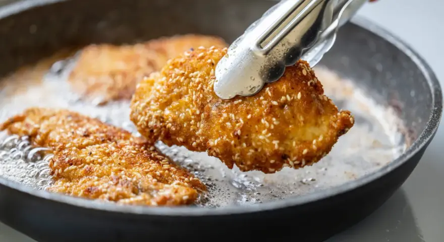 Une escalope de dinde panée aux graines de sésame est retournée dans une poêle avec de l'huile chaude.