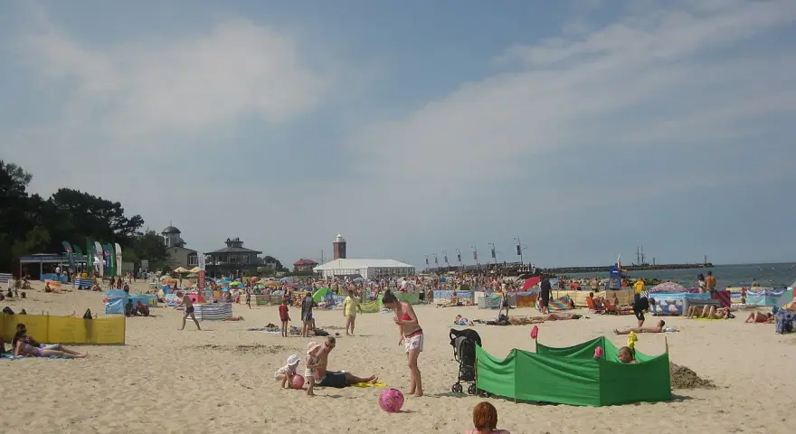 Słoneczna plaża w Darłowie tętni życiem. Ludzie opalają się, bawią z dziećmi i odpoczywają pod kolorowymi parawanami.