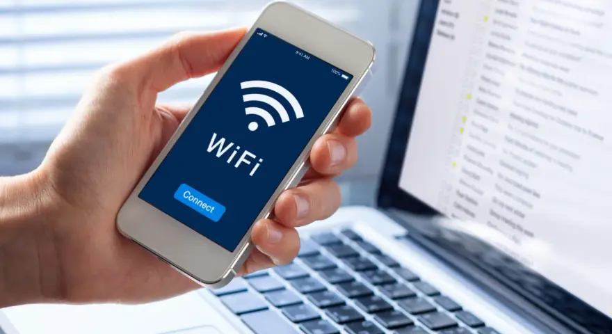 Dłoń trzyma smartfon z ikoną WiFi i napisem "Connect". Obok widać laptopa z listą e-maili, sugerującą pracę zdalną i dostęp do sieci, być może przez **ssid** domowy.
