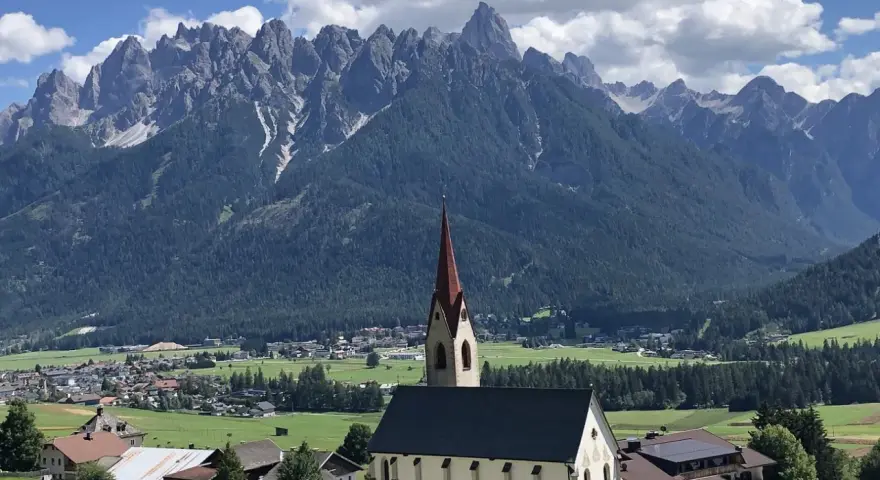 Chiesa con campanile rosso in primo piano, sullo sfondo le maestose vette delle Dolomiti e i **paesi più belli della Val Pusteria**.