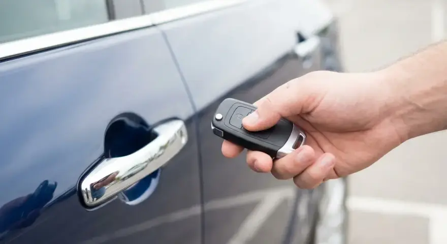 Dłoń trzyma kluczyk keyless, zbliżając go do klamki ciemnoniebieskiego samochodu.
