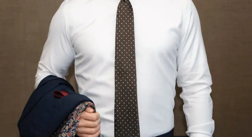 Elegancki mężczyzna w białej koszuli i modnym krawacie męskim w kropki, trzymający granatową marynarkę z wzorzystą podszewką.