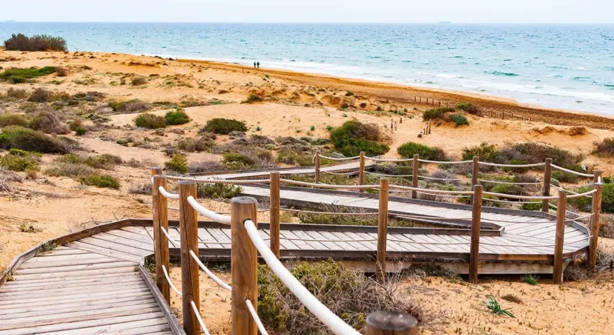 Pasarela de madera serpentea por la duna, invitando a explorar la ruta Calblanque hacia el mar azul.