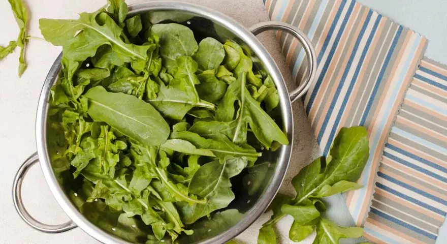Rukola w durszlaku, idealna do przygotowania jadłospisu tygodniowego dieta FODMAP. Świeże liście na tle kolorowych tkanin.
