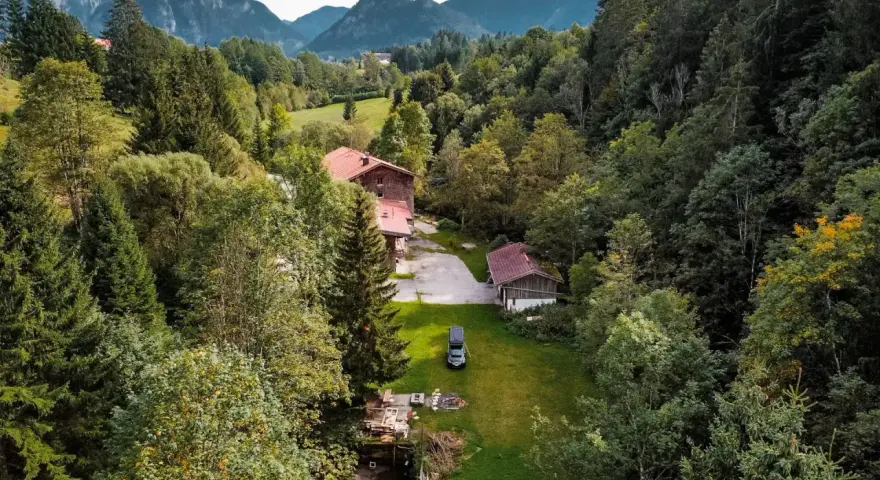 Idyllisches **Camping in den Bergen**: Ein Wohnmobil steht auf einer grünen Wiese neben einem Bach, umgeben von dichten Wäldern und traditionellen Holzhäusern.