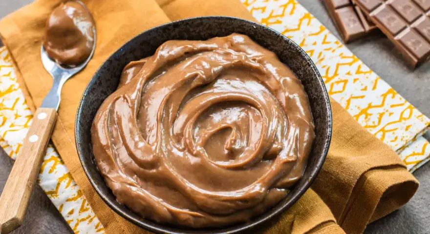 Una ciotola piena di deliziosa crema pasticcera al cioccolato, con una cucchiaiata pronta da gustare. Accanto, una tavoletta di cioccolato.