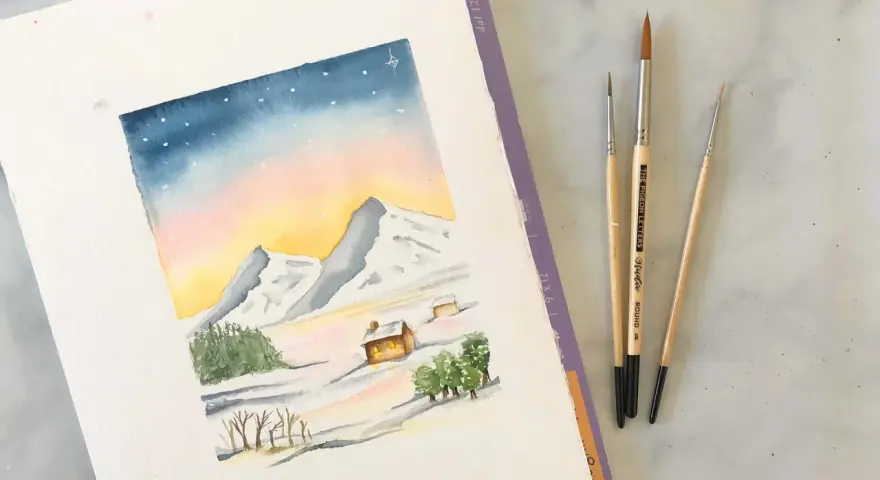 Une aquarelle montagne facile dépeint un paysage hivernal doux avec des montagnes enneigées, des chalets chaleureux et un ciel étoilé. Trois pinceaux sont posés à côté.