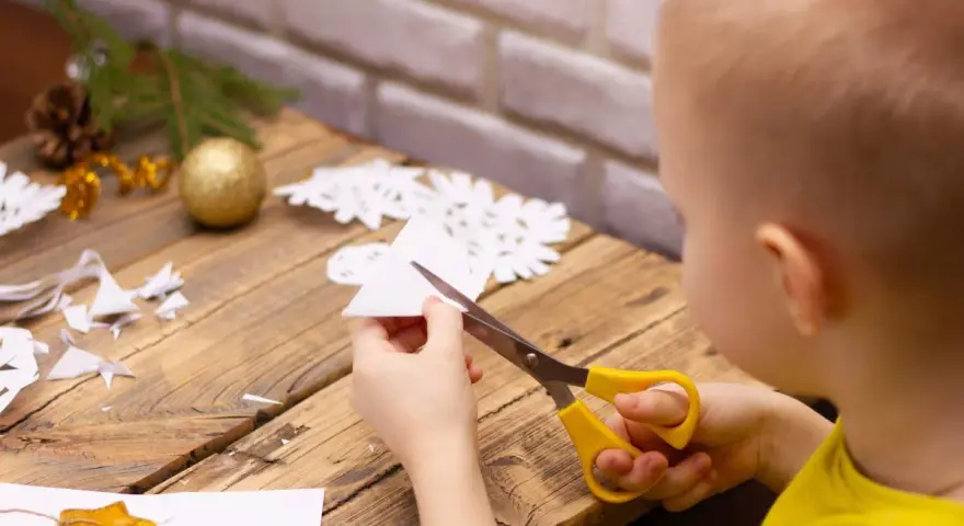 Małe rączki wycinają śnieżynkę z papieru. Na stole leżą gotowe ozdoby i gałązka choinki.