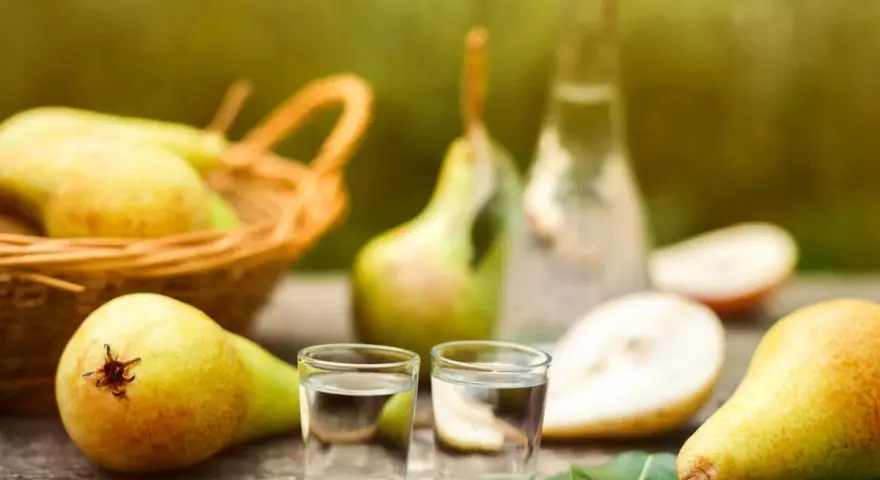 Frische Birnen und zwei Gläschen Schnaps auf Holztisch. Ein Korb voller Birnen im Hintergrund. Perfekt für ein Birnenschnaps Rezept.