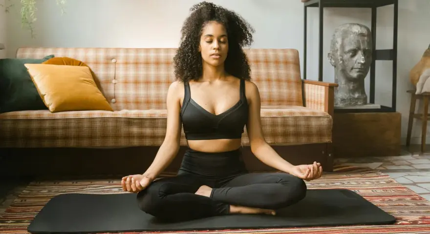 Frau in schwarzer Sportkleidung praktiziert Meditation für tiefe Entspannung auf einer Matte vor einem karierten Sofa.