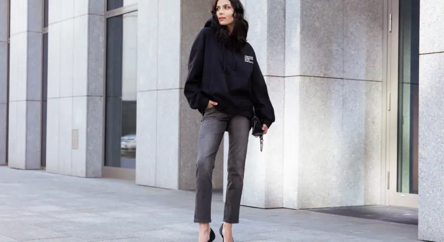 Kobieta w czarnej bluzie z kapturem i szarych jeansach. Luźny krój (loose fit) bluzy podkreśla swobodny styl.