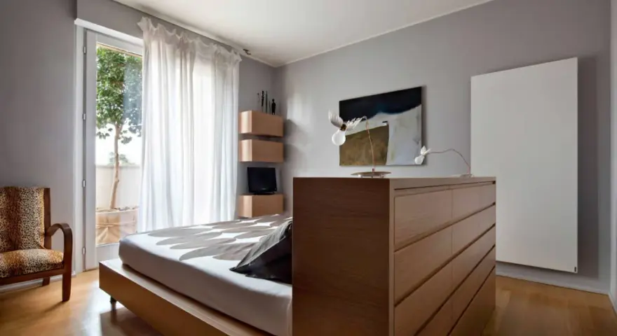 Un moderno **letto in mezzo alla stanza**, con un copriletto a motivi floreali, accanto a una poltrona leopardata e una cassettiera in legno.