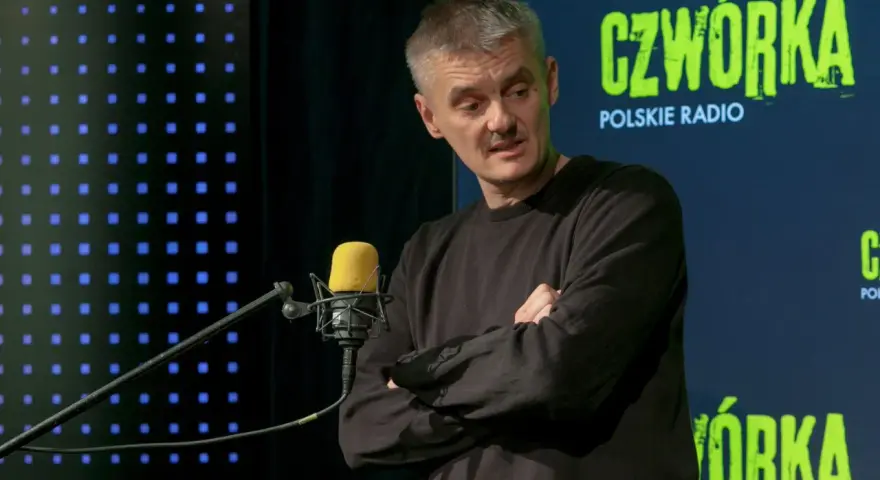 Jacek Sienkiewicz w studiu Polskiego Radia, przed mikrofonem, z rękami skrzyżowanymi na piersi.
