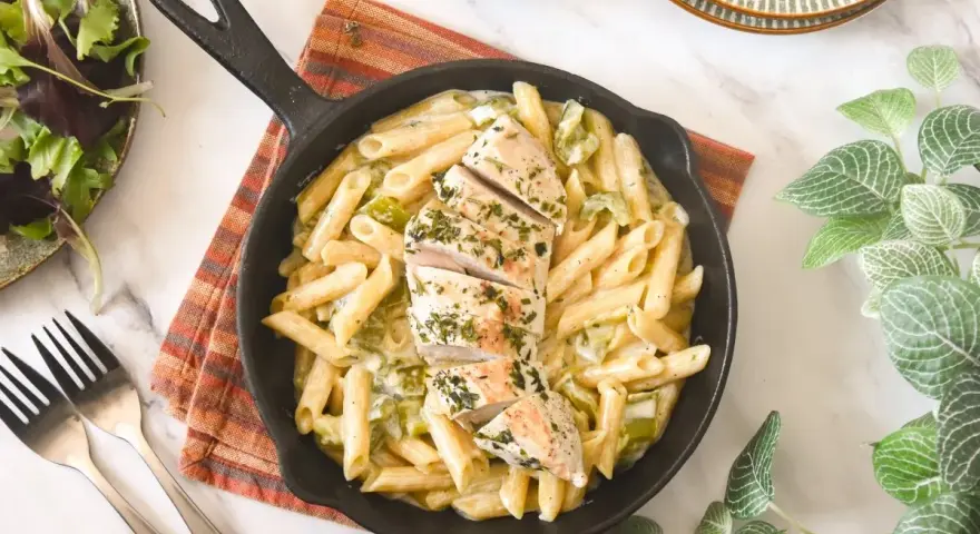 Deliciosa pasta con pollo en sartén, cubierta con hierbas frescas. Acompañada de una ensalada verde y tenedores.