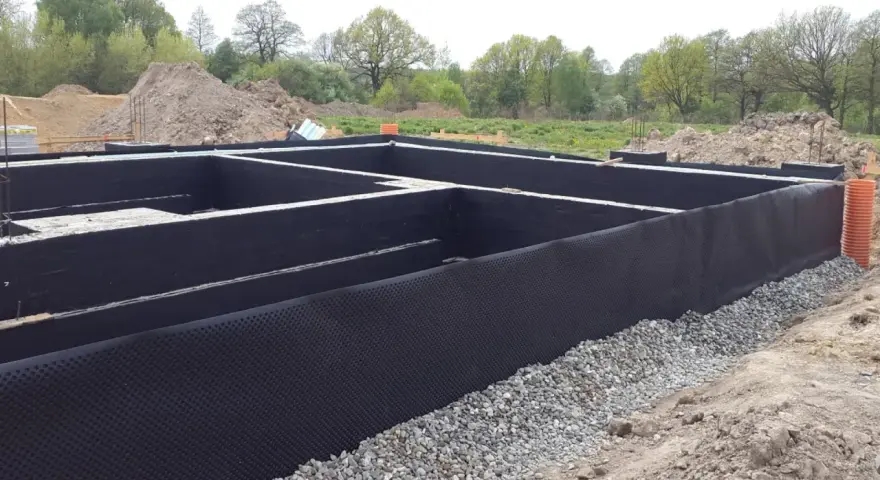 Fundament domu w trakcie budowy, z czarną membraną i otoczakami. Trwa ocieplenie fundamentów, przygotowanie do dalszych prac.