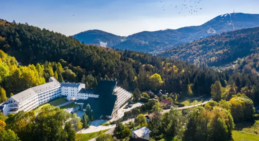 Malowniczy hotel przy stoku narciarskim w Polsce, otoczony jesiennym lasem. W oddali góry i niebo z ptakami.