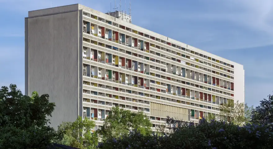 Długi budynek mieszkalny w stylu modernizm, z rzędami balkonów w kolorowych obramowaniach, otoczony zielenią.