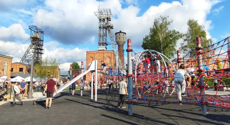 Rybnik atrakcje: plac zabaw dla dzieci z linowymi konstrukcjami i zjeżdżalnią, w tle wieże szybowe kopalni.